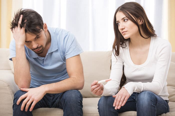 Upset woman is looking in pregnancy test. Frustrated man is sitting next to her.Portrait of frustrated couple are sitting on couch and are quarreling with each other.